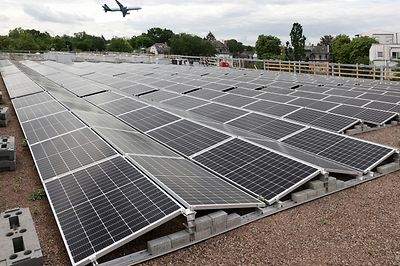 © Mainova Dach mit vielen Solarpaneelen, im Hintergrund fliegt ein Flugzeug über grüne Landschaft.