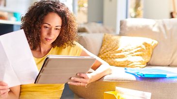 Frau sitzt vor Couch und schaut auf Tablet mit Dokument in der Hand