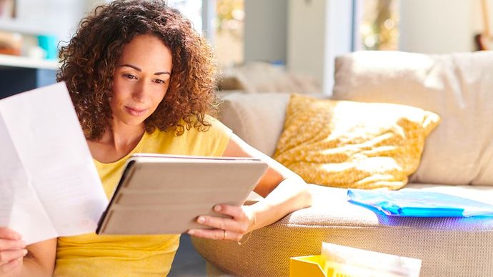 Frau sitzt vor Couch und schaut auf Tablet mit Dokument in der Hand