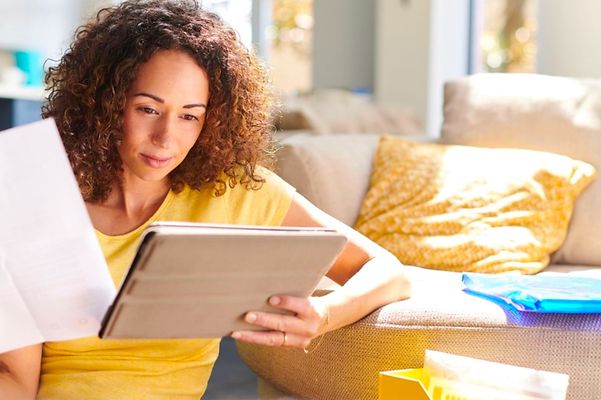 Frau sitzt vor Couch und schaut auf Tablet mit Dokument in der Hand