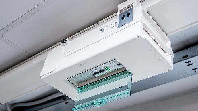 Ein weißer Verteilerkasten mit grünem, teils geöffnetem Deckel, montiert an der Decke. Sichtbare Schalter und Verkabelung.