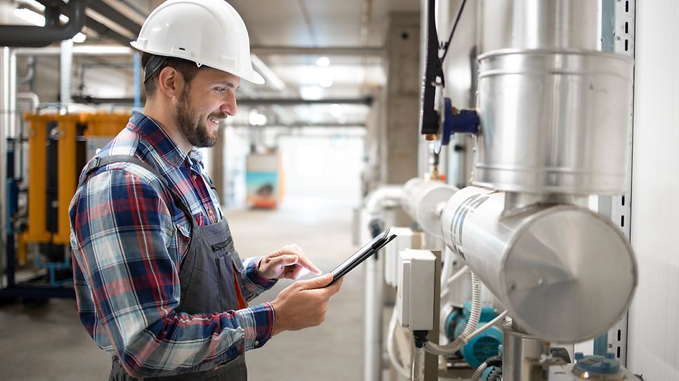© istockphoto.com/Smederevac Installateur im Heizungraum steht an den Leitungen und prüft mit dem Tablett die Werte