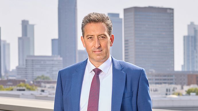 Mann im blauen Anzug mit roter Krawatte blickt ernst in die Kamera. Stadthintergrund mit Wolkenkratzern.