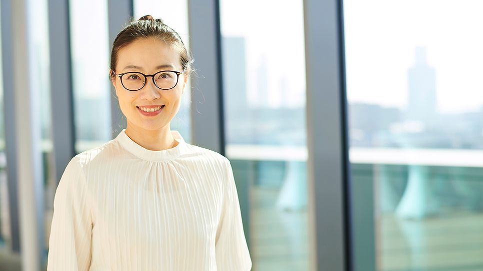 © Mainova Frau mit Brille lächelt in hellem Raum, weißes Oberteil, Fenster mit Stadtblick dahinter.
