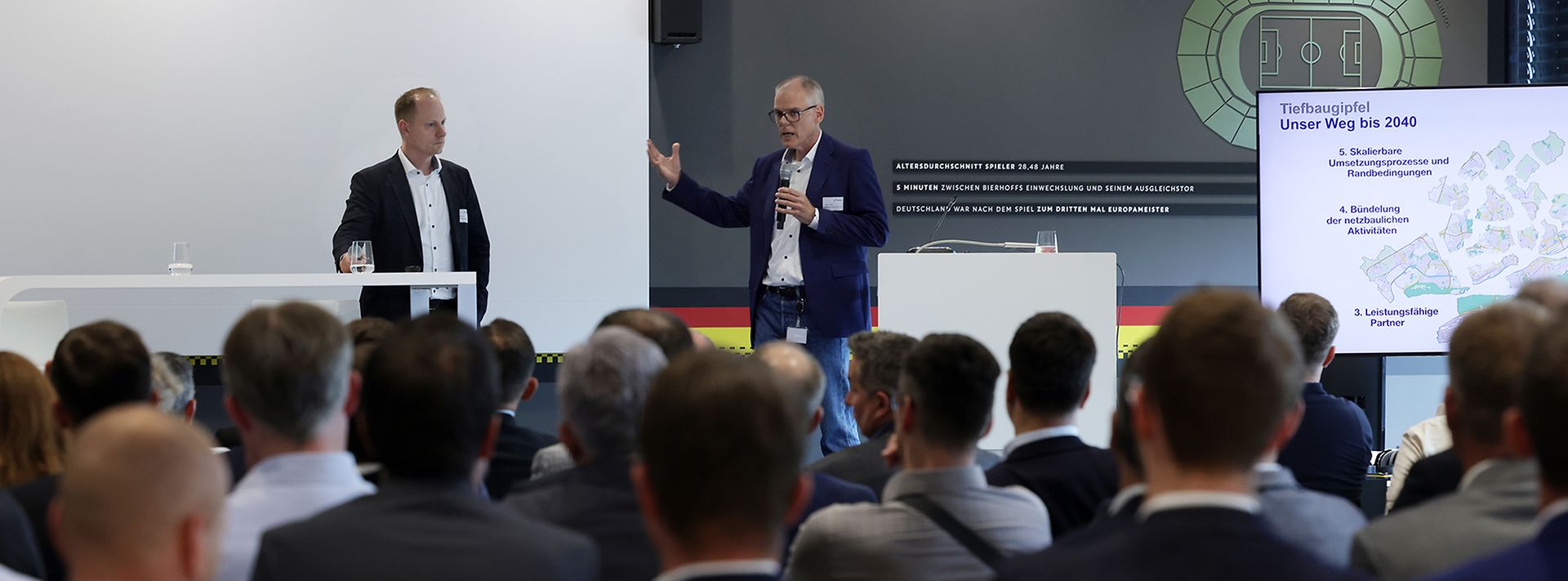 © Mainova Ein Redner spricht auf einer Konferenz, während das Publikum zuhört. Eine Präsentation zum "Tiefbaugipfel" wird angezeigt.