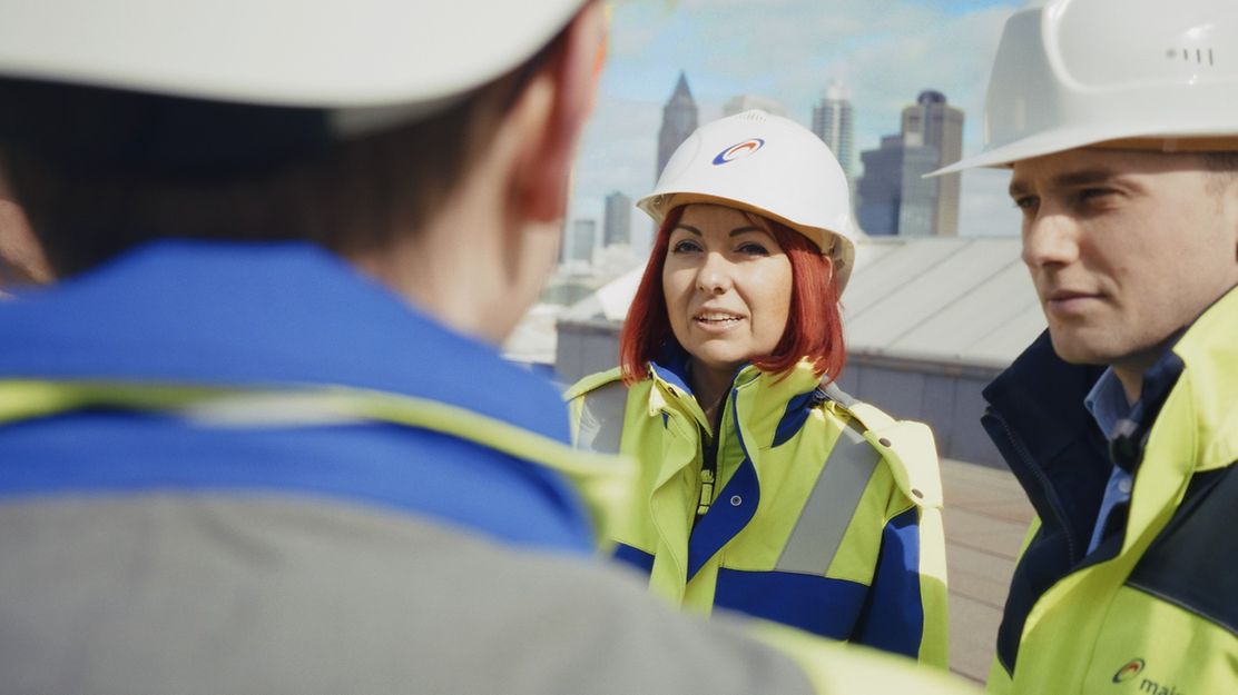 © Mainova Eine Frau mit rotem Haar und Schutzhelm trägt eine gelbe und blaue Jacke in einem industriellen Gebäude.