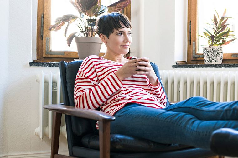 © AdobeStock/Westend61 Eine Frau in einem gestreiften Pullover sitzt entspannt in einem Sessel, hält eine Tasse und blickt aus dem Fenster.