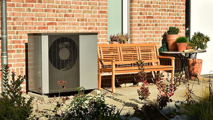 Wärmepumpe vor rot verziegelter Hauswand, daneben Sitzbank und Tisch mit Topfpflanzen.