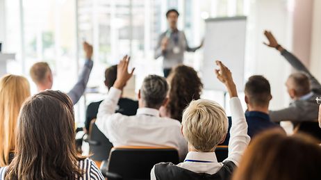 © iStock.com/skynesher Personengruppe in einem Raum versammelt, stellt Fragen an eine Person die mit einem Flipchart eine Präsentation hält