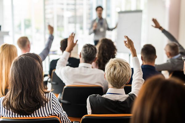 Personengruppe in einem Raum versammelt, stellt Fragen an eine Person die mit einem Flipchart eine Präsentation hält 