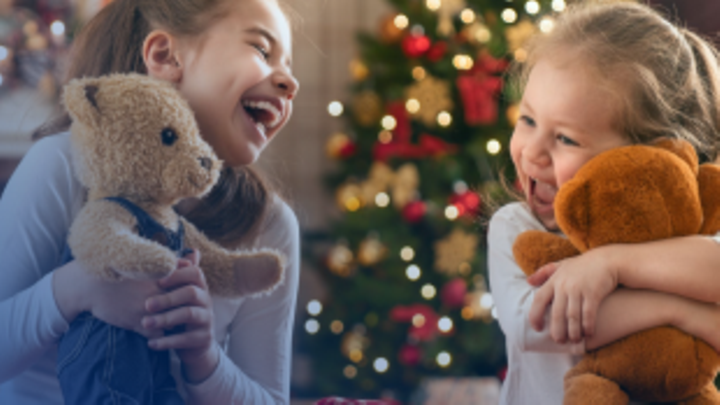 Zwei kleine Mädchen spielen und kuscheln mit ihren Teddybären vor einem Weihnachtsbaum