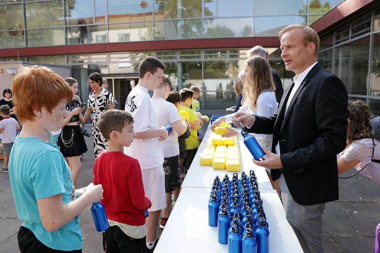 © Heiko Rhode Ein langer Tisch, links stehen Kinder und rechts stehen Erwachsene, Kinder bekommen blaue Trinkflaschen und gelbe Brotboxen