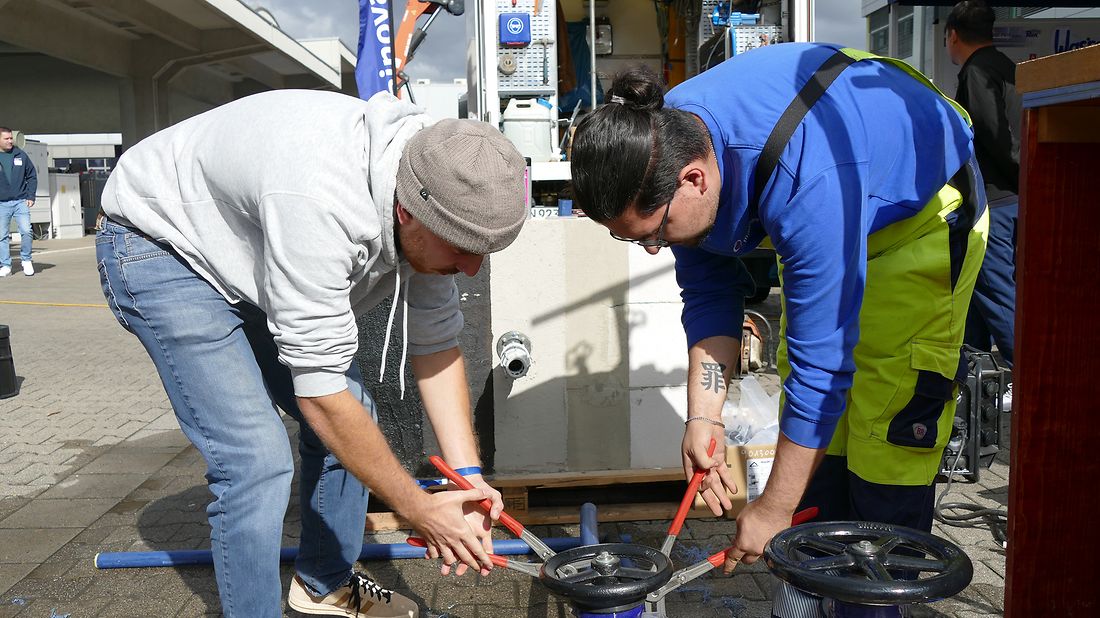 Zwei Männer arbeiten im Freien mit Rohrzangen an einer großen blauen Wasserleitung. Ein Bagger steht im Hintergrund.