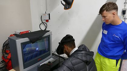 Junger Mann mit VR-Headset nutzt ein Schweiß-Simulationsgerät. Ein Auszubildender in blauer Kleidung beobachtet ihn.