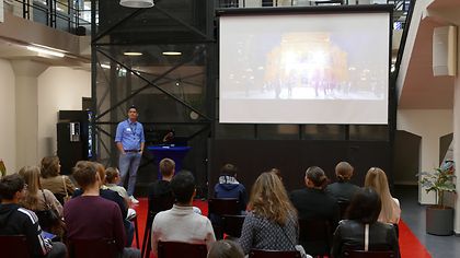 Mann hält eine Präsentation vor einer Gruppe von Zuhörern. Auf der Leinwand ist ein beleuchtetes Gebäude bei Nacht zu sehen.