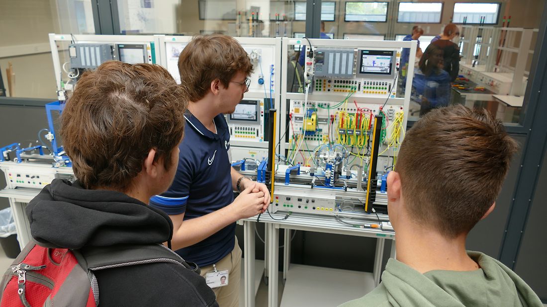 Mann erklärt zwei Jugendlichen eine technische Anlage mit vielen Kabeln und Bildschirmen. Sie hören aufmerksam zu.