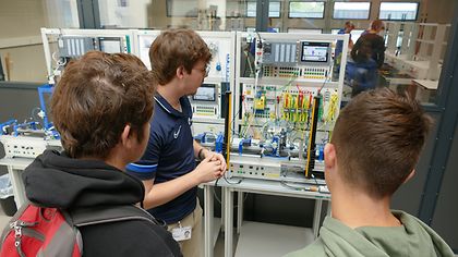 Mann erklärt zwei Jugendlichen eine technische Anlage mit vielen Kabeln und Bildschirmen. Sie hören aufmerksam zu.