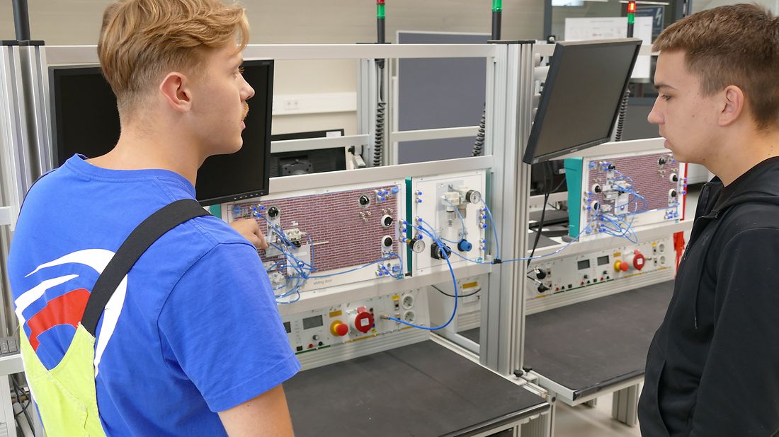 Zwei junge Männer stehen vor einer technischen Versuchsanlage mit Sensoren und Bildschirmen. Einer erklärt dem anderen etwas.