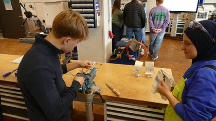 Junge feilt Metall in einer Werkstatt, junge Frau mit Kopftuch wischt Tisch ab. Im Hintergrund arbeiten weitere Personen.