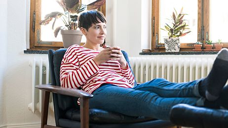 Eine Frau in einem gestreiften Pullover sitzt entspannt in einem Sessel, hält eine Tasse und blickt aus dem Fenster.