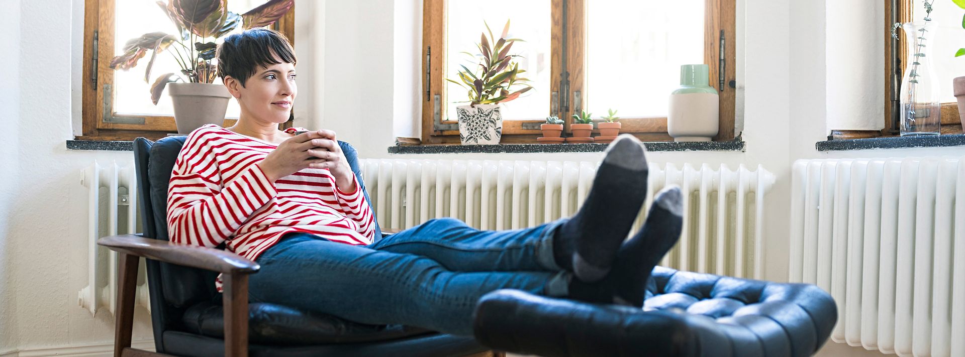 Eine Frau in einem gestreiften Pullover sitzt entspannt in einem Sessel, hält eine Tasse und blickt aus dem Fenster.