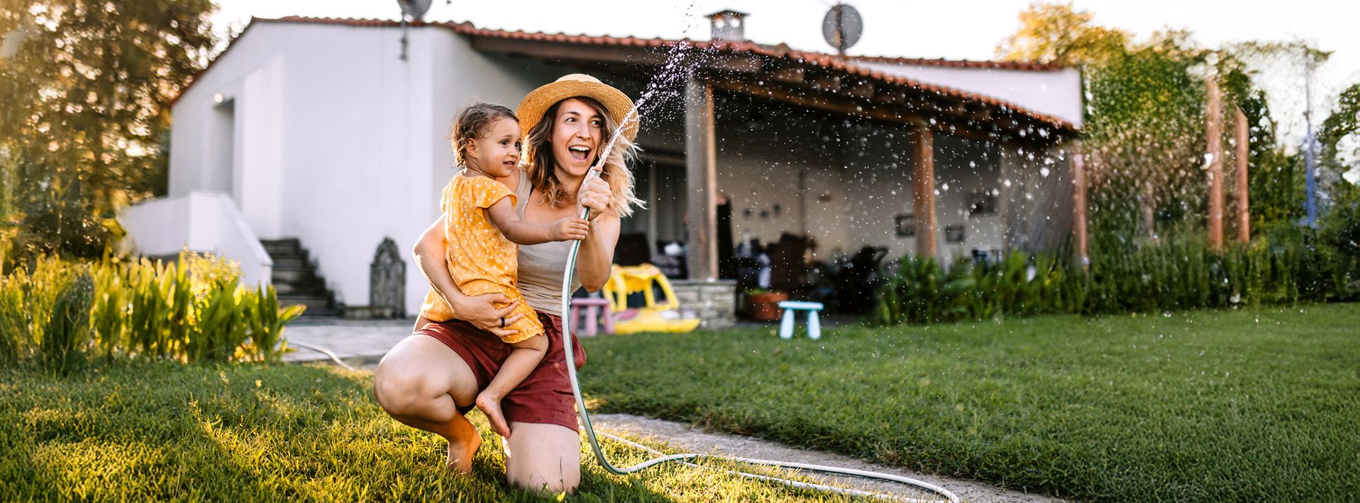 Eine Mutter und ihre kleine Tochter spielen im Garten mit einem Gartenschlauch.