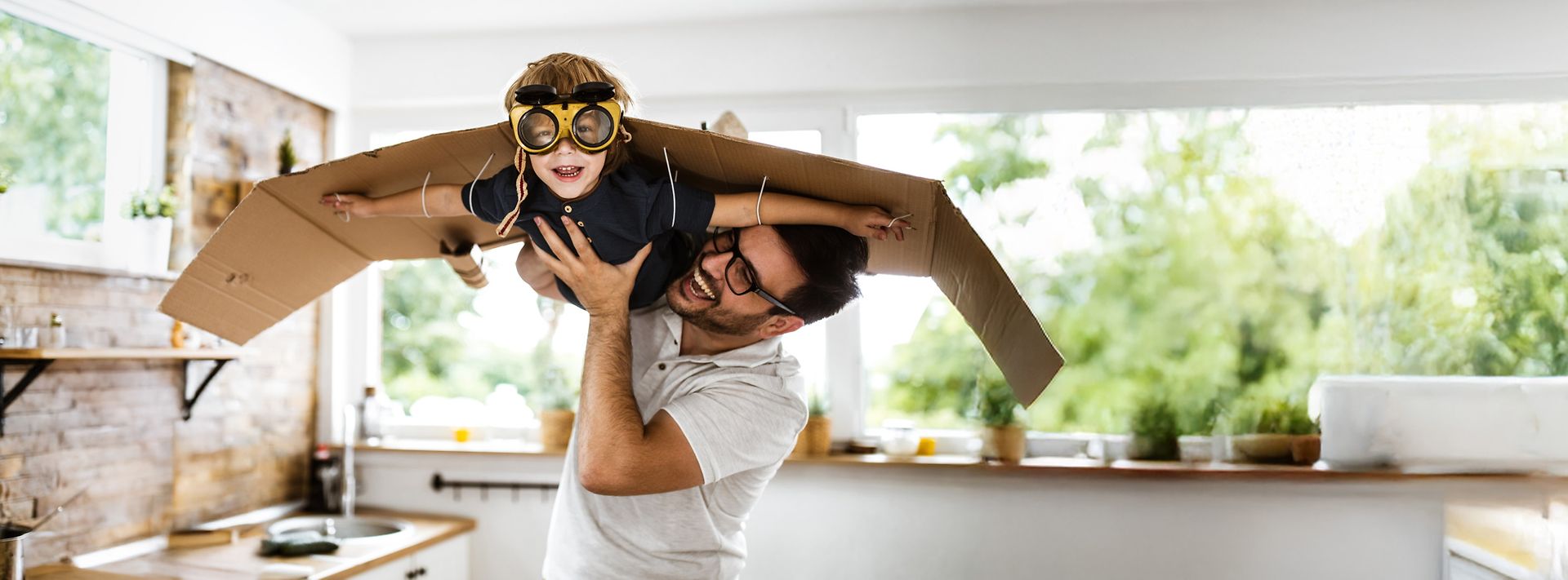 © skynesher Ein Vater hält seinen Sohn in die Luft, welcher mit Fliegerbrille und angeklebten Pappflügeln durch das Zimmer fliegt.