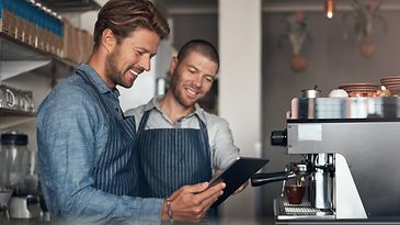 Zwei Mitarbeiter in einem Café stehen vor einer Kaffeemaschine und schauen gemeinsam auf ein Tablet.