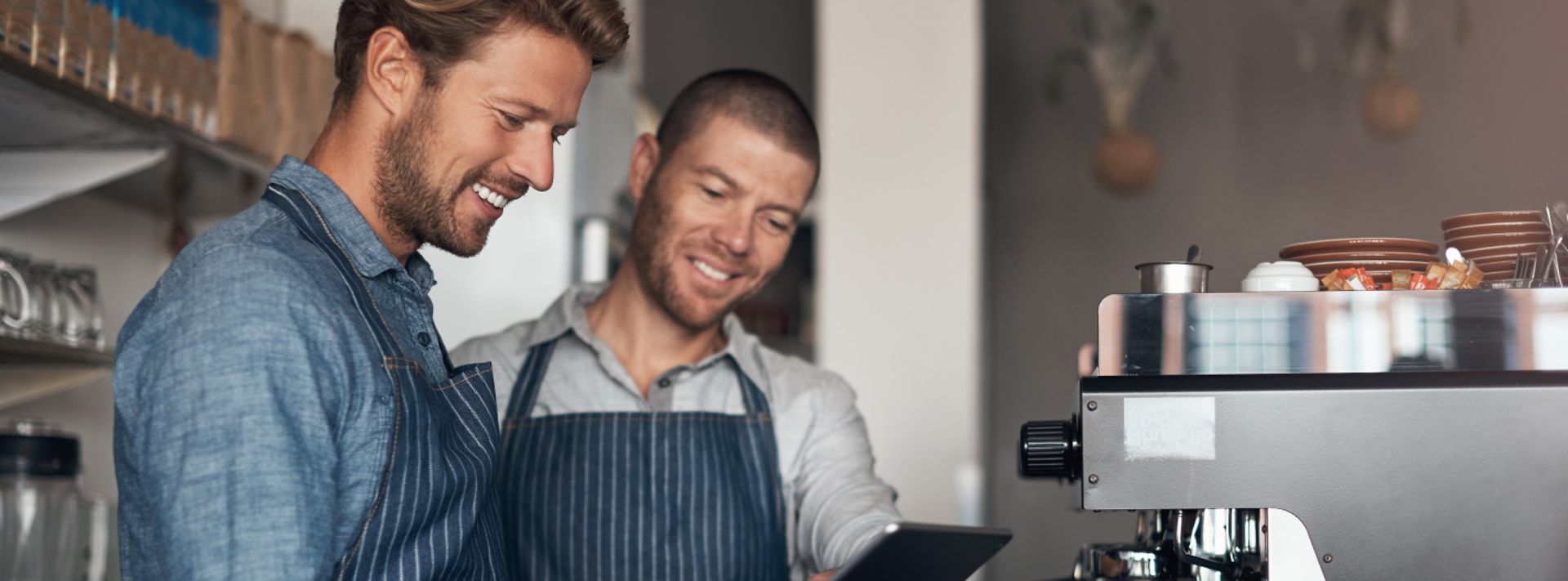Zwei Mitarbeiter in einem Café stehen vor einer Kaffeemaschine und schauen gemeinsam auf ein Tablet.