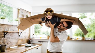 Ein Vater hält seinen Sohn in die Luft, welcher mit Fliegerbrille und angeklebten Pappflügeln durch das Zimmer fliegt.
