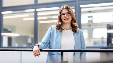 © Mainova/DennisMöbus Eine junge Frau mit Brille und welligem Haar lehnt lächelnd am Geländer in einem modernen Büro mit Glaswänden.