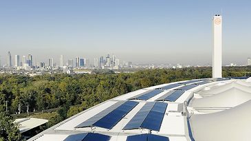 © studio fiftynine Solarmodule auf einem geschwungenen Dach, dahinter Frankfurts Skyline und viel Grün bei klarem Himmel im Panorama.