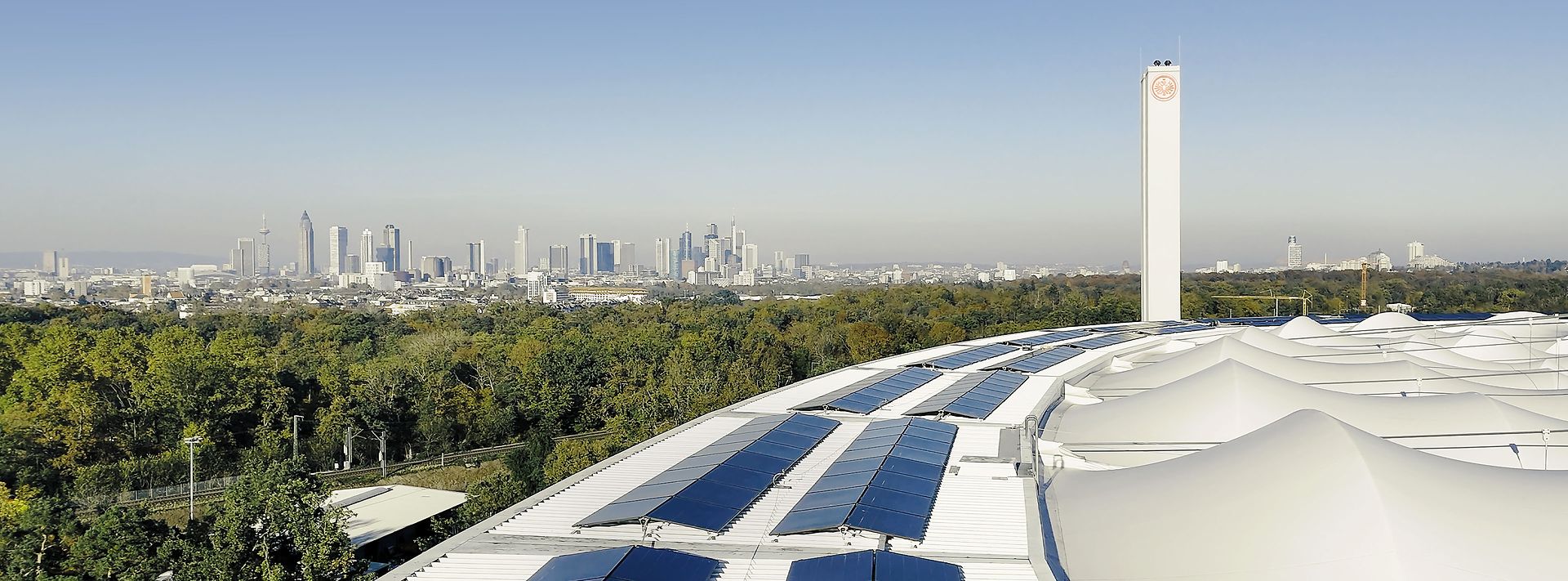 © studio fiftynine Solarmodule auf einem geschwungenen Dach, dahinter Frankfurts Skyline und viel Grün bei klarem Himmel im Panorama.
