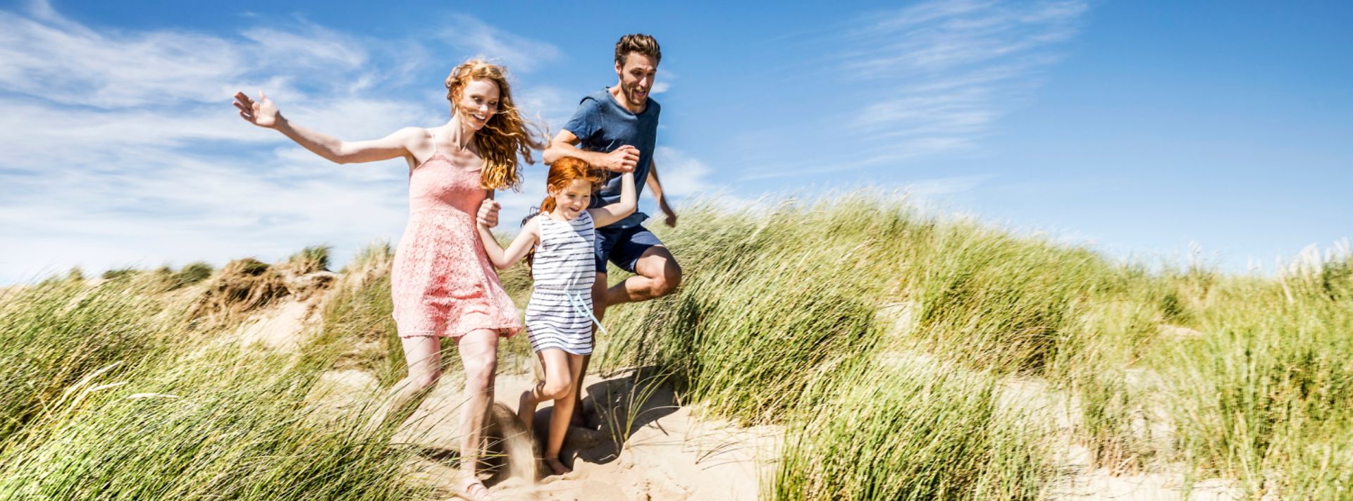 © Westend61 Eine Familie läuft durch Sanddünen mit Gras unter einem klaren blauen Himmel.