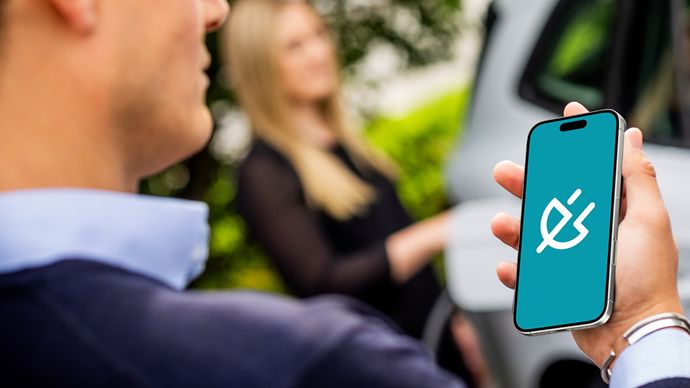 Eine Person hält ein Smartphone, trägt ein dunkelblaues Oberteil. Im Hintergrund steht eine Person vor einem weißen Auto.