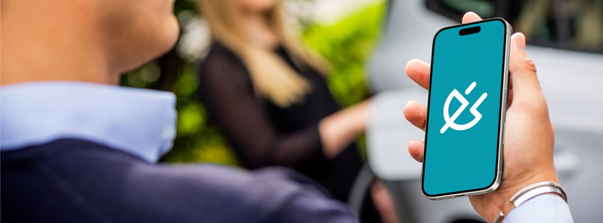 © Dennis Möbus Eine Person hält ein Smartphone, trägt ein dunkelblaues Oberteil. Im Hintergrund steht eine Person vor einem weißen Auto.