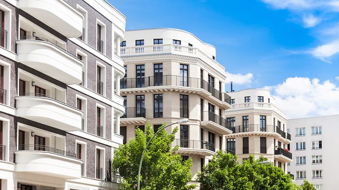 © Tiberius Gracchus - stock.adobe.com Moderne Wohngebäude mit Balkonen und großen Fenstern, umgeben von Bäumen unter einem blauen Himmel mit weißen Wolken.