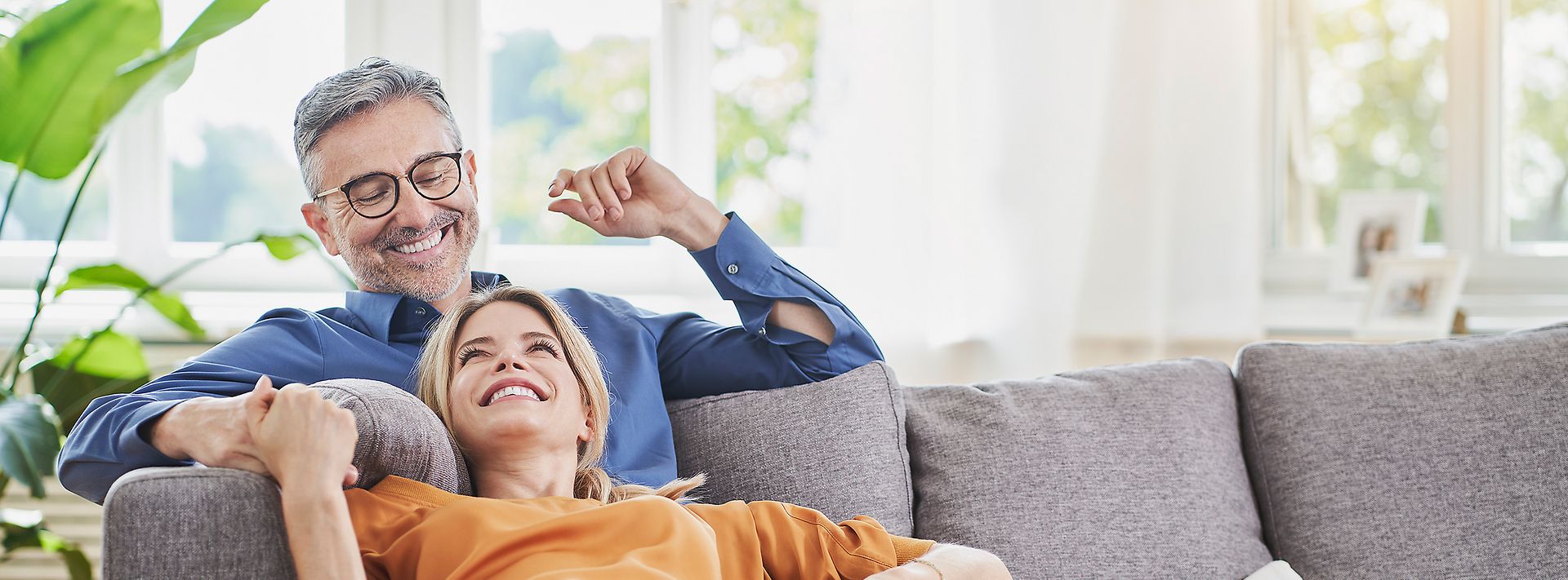 Ein Paar sitzt lachend und entspannt auf einem Sofa in einem hellen Wohnzimmer mit großen Fenstern und Pflanzen.