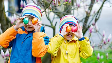 Zwei Kinder halten bunte Ostereier vor das Gesicht und stehen in farbigen Jacken vor einem blühenden Magnolienbaum.