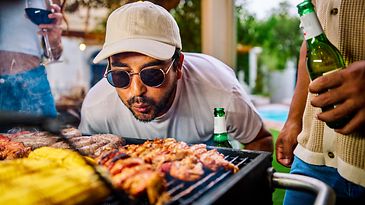 Mann mit Sonnenbrille pustet auf einen Grill mit Grillgut