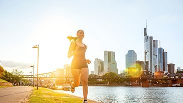 Frau joggt lächelnd am Main vor der Frankfurter Skyline