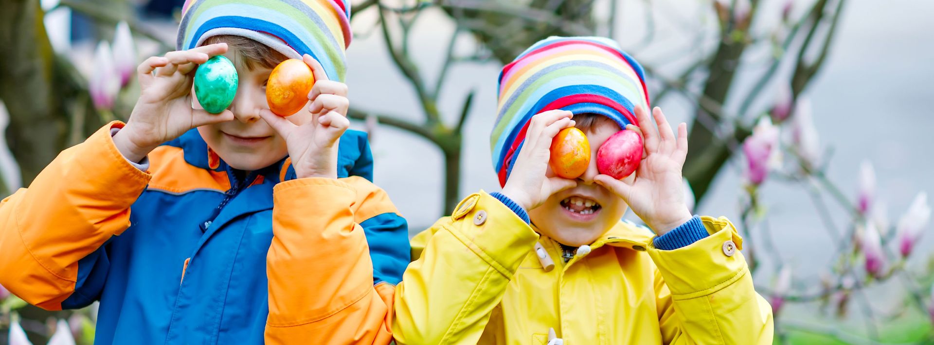 Zwei Kinder halten bunte Ostereier vor das Gesicht und stehen in farbigen Jacken vor einem blühenden Magnolienbaum.