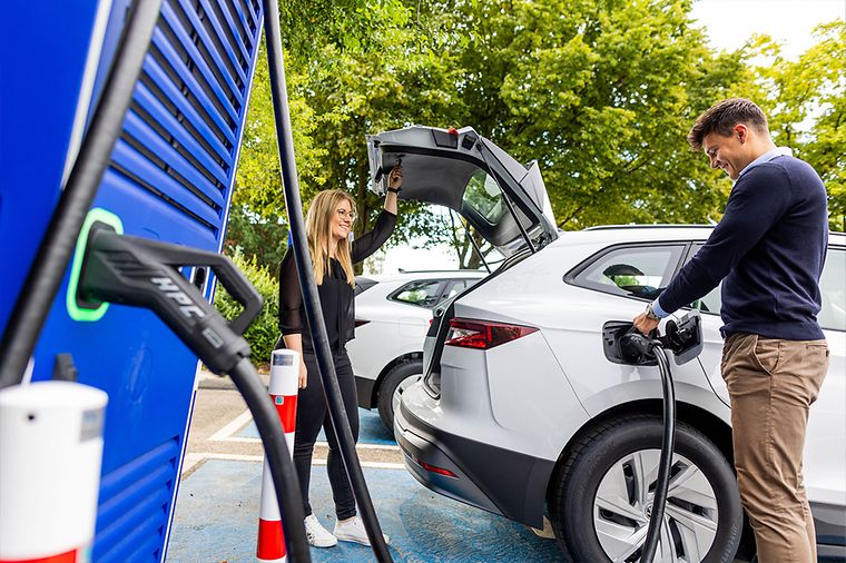 © Dennis Möbus Frau und Mann mit weißem E-Auto an Ladesäule, Mann lädt Auto