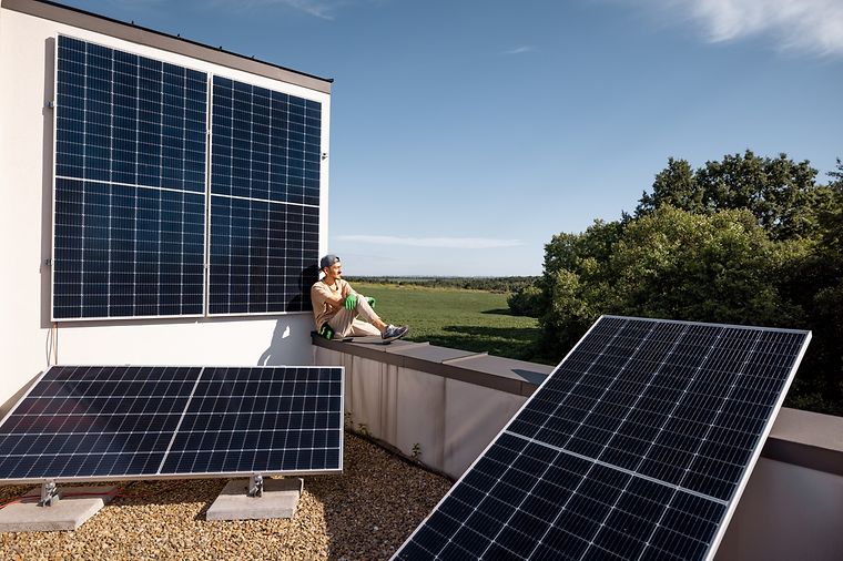 Junger Mann mit Arbeitshandschuhen sitzt auf einem Balkon mit drei großen Solarpanelen