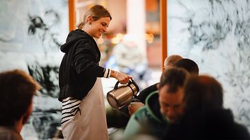 © NoiCrew Frau schenkt Mann Kaffee aus einer Kanne ein.