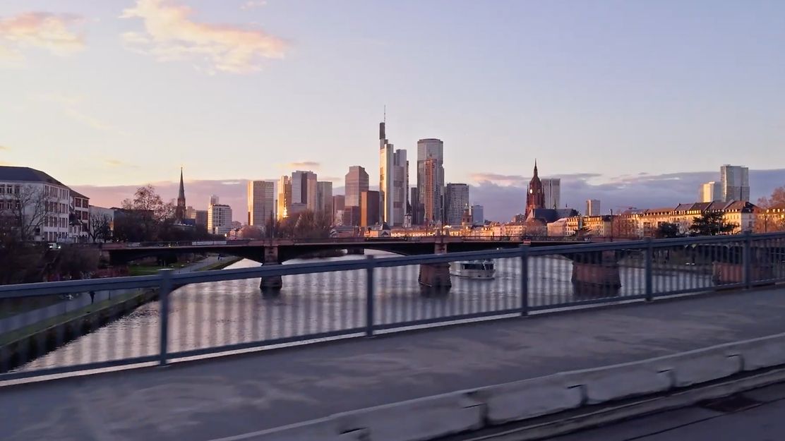 Skyline von Frankfurt bei Sonnenuntergang, mit Mainbrücke, Boot und reflektierenden Hochhäusern.