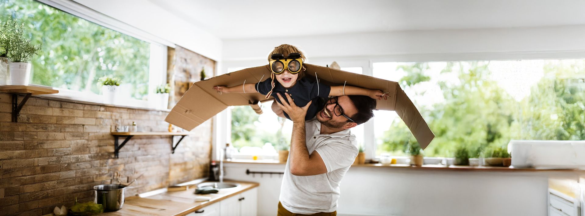 Ein Vater hält seinen Sohn in die Luft, welcher mit Fliegerbrille und angeklebten Pappflügeln durch das Zimmer fliegt.