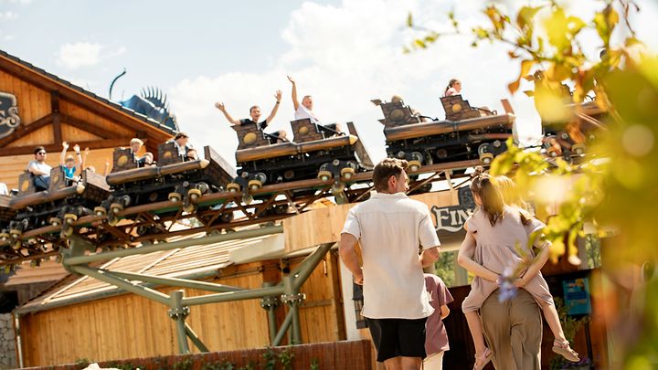 Personen gehen an einer Holzachterbahn vorbei, während Wagen über ihnen entlangfahren.