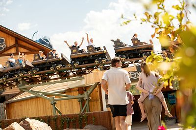 Personen gehen an einer Holzachterbahn vorbei, während Wagen über ihnen entlangfahren.