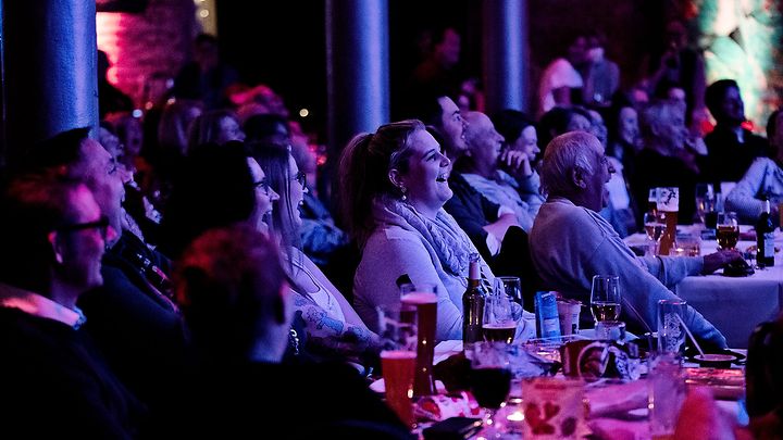 Publikum sitzt an Tischen in farbig beleuchtetem Saal und blickt geschlossen Richtung Bühne.